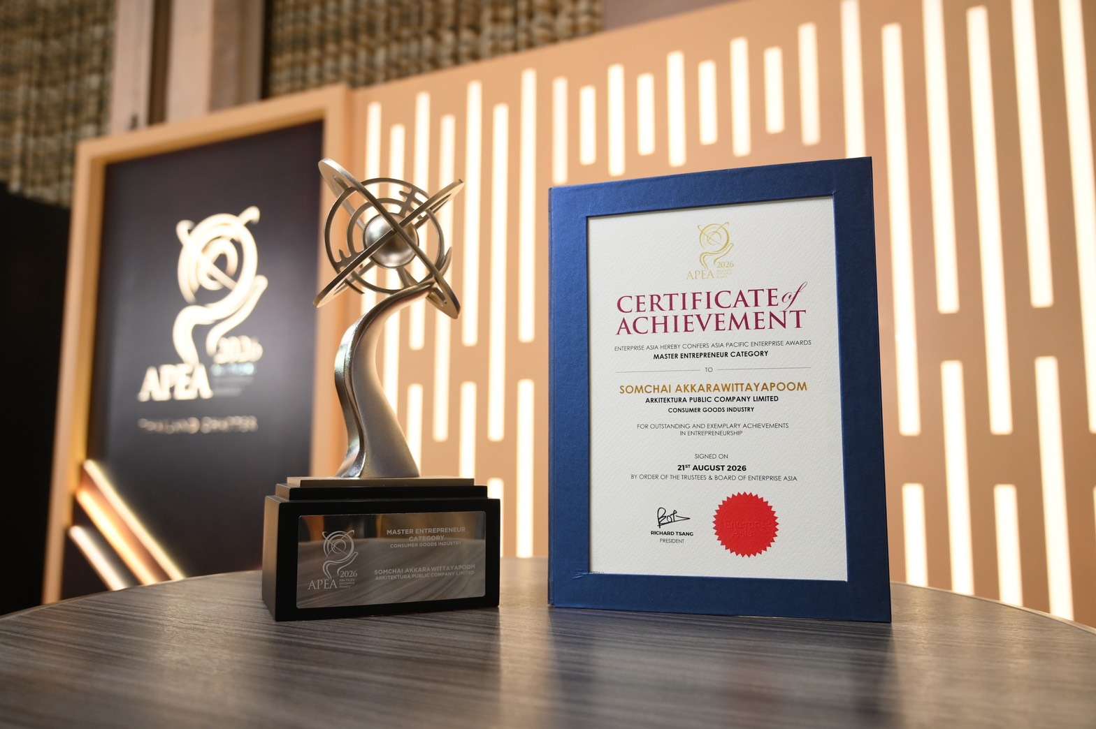 ภาพถ่ายเจาะจงรายละเอียดของถ้วยรางวัลดีไซน์โดดเด่นและประกาศนียบัตร Master Entrepreneur Award จากงาน APEA 2026 ที่ระบุชื่อนายสมชัย อัครวิทยาภูมิ แห่งบริษัท อาร์คิเทคทูรา จำกัด (มหาชน)