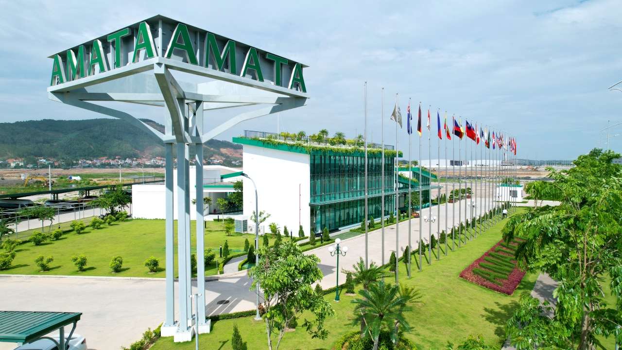 ป้ายสัญลักษณ์ตัวอักษร AMATA สีเขียวขนาดใหญ่ตั้งอยู่บริเวณทางเข้านิคมอุตสาหกรรมอมตะในประเทศเวียดนาม พร้อมด้วยเสาธงชาติต่างๆ ที่เรียงรายอยู่ด้านหน้าอาคารสำนักงานกระจก