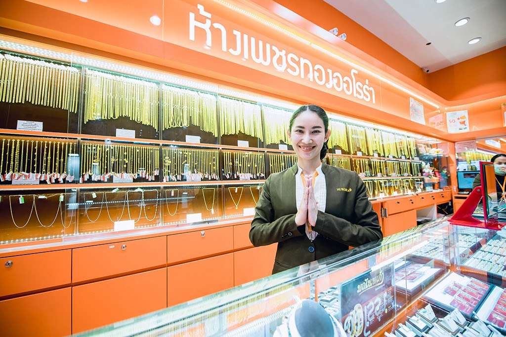 ภาพพนักงานหญิงของห้างเพชรทองออโรร่า สวมชุดยูนิฟอร์มสีเขียว ยืนพนมมือไหว้ต้อนรับลูกค้าด้วยรอยยิ้มสดใส โดยมีฉากหลังเป็นตู้กระจกแสดงสินค้าทองคำ