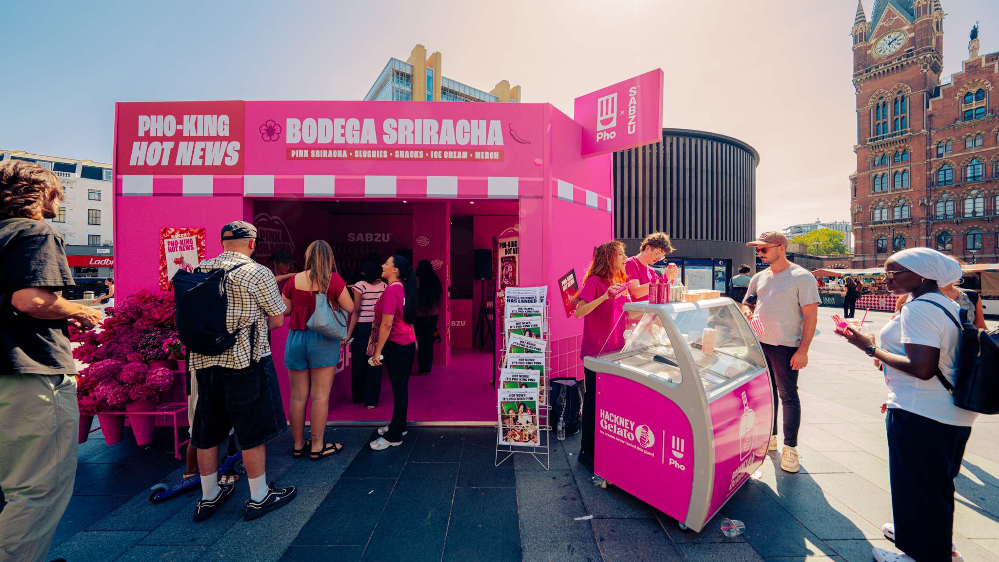 ภาพบรรยากาศบูธกิจกรรม Bodega Sriracha สีชมพูสดใส ตั้งอยู่บริเวณลานหน้าสถานีรถไฟ King’s Cross กรุงลอนดอน โดยมีผู้คนให้ความสนใจร่วมกิจกรรม พร้อมจุดบริการไอศกรีม Hackney Gelato และชั้นวางหนังสือพิมพ์จำลอง
