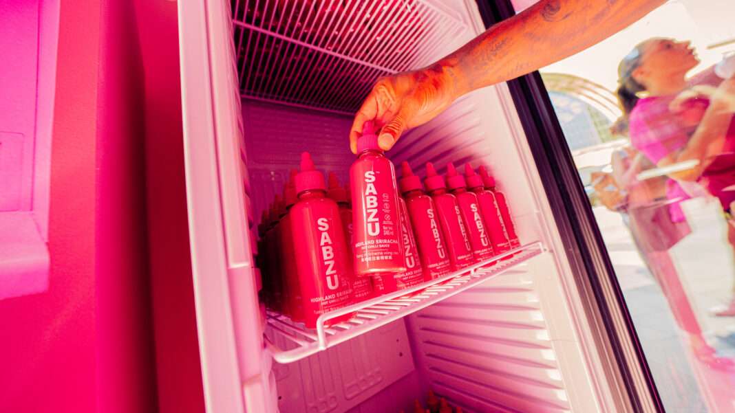 SABZU-Pink-Sriracha-Bottle-NRF-Premium-Sauce ภาพมือของลูกค้ากำลังหยิบขวดซอสพริก Pink Sriracha แบรนด์ SABZU จากตู้แช่เย็นสีชมพู สะท้อนภาพลักษณ์ผลิตภัณฑ์เครื่องปรุงรสพรีเมียมภายใต้เครือ NRF ที่โดดเด่นและดึงดูดสายตา