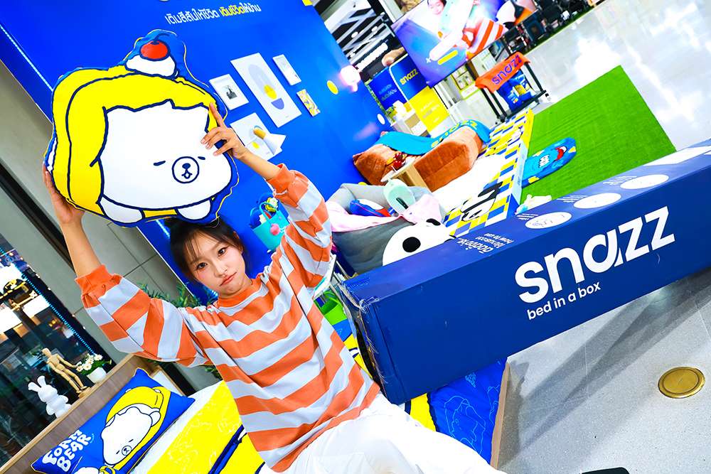หญิงสาววัยรุ่นสวมเสื้อลายขวางกำลังถือหมอนอิงลวดลายน่ารักจากแบรนด์ SNAZZ โดยมีสินค้าไฮไลต์คือ SNAZZ Bed in a Box หรือที่นอนกล่องสูญญากาศวางอยู่ด้านข้าง
