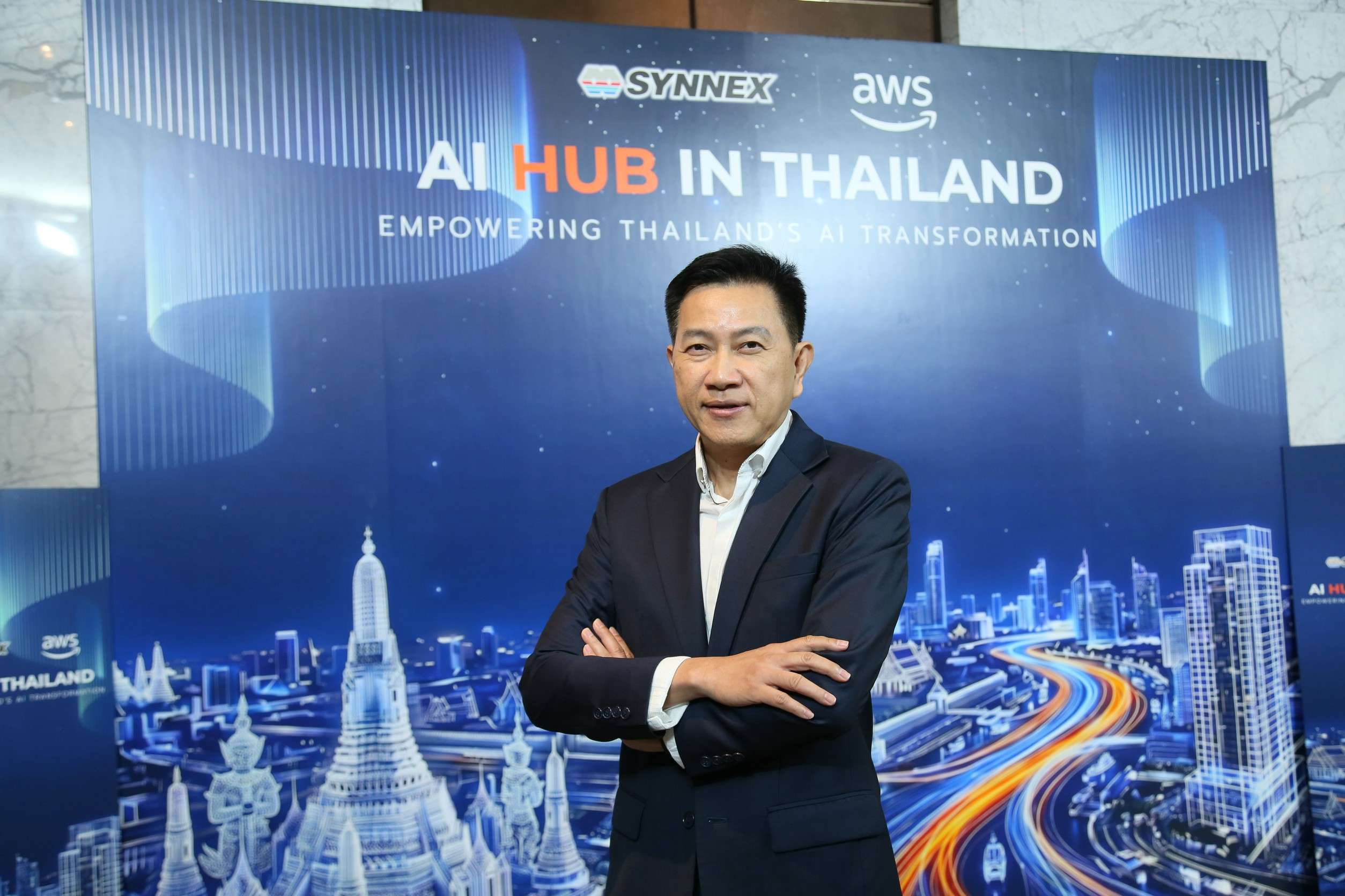 ภาพคุณวัตสัน ถิรภัทรพงศ์ Country Manager ของบริษัท อะเมซอน เว็บ เซอร์วิสเซส (ประเทศไทย) จำกัด (AWS) สวมชุดสูทสีกรมท่า ยืนกอดอกด้วยความมั่นใจ ถ่ายภาพบริเวณหน้าแบคดรอปงานเปิดตัว AI HUB IN THAILAND ภายใต้ความร่วมมือของ SYNNEX และ AWS