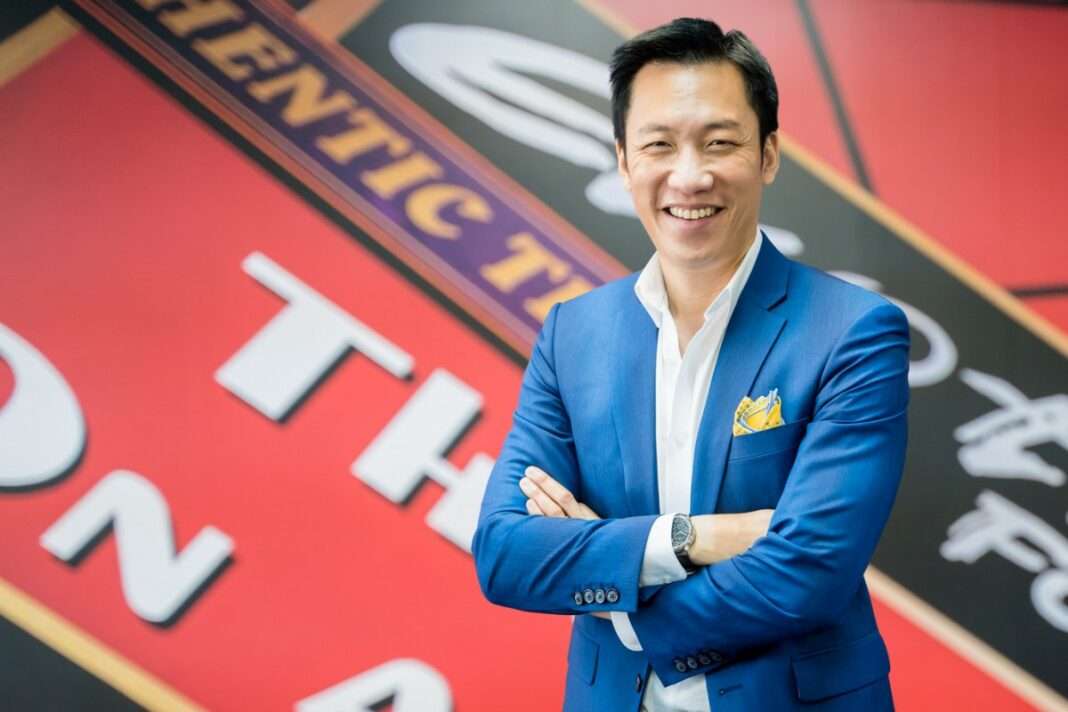 jittiporn-jantarach-ceo-exotic-food-xo-q3-2026-outlook ภาพนายจิตติพร จันทรัช ประธานเจ้าหน้าที่บริหาร บริษัท เอ็กโซติค ฟู้ด จำกัด (มหาชน) หรือ XO สวมสูทสีน้ำเงิน ยืนกอดอกยิ้มแย้ม โดยมีฉากหลังเป็นป้ายแบรนด์ซอสพริกศรีราชา เพื่อนำเสนอทิศทางธุรกิจและผลประกอบการ