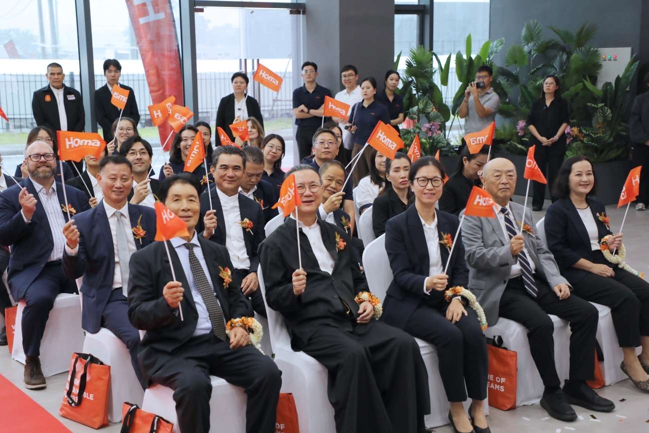ภาพบรรยากาศผู้เข้าร่วมงานและคณะผู้บริหาร รวมถึงนายวิกรม กรมดิษฐ์ และนายไมเคิล เหยา ที่นั่งอยู่แถวหน้าสุด กำลังยิ้มแย้มและโบกธงสีส้มสัญลักษณ์ Homa เพื่อเฉลิมฉลองความร่วมมือ