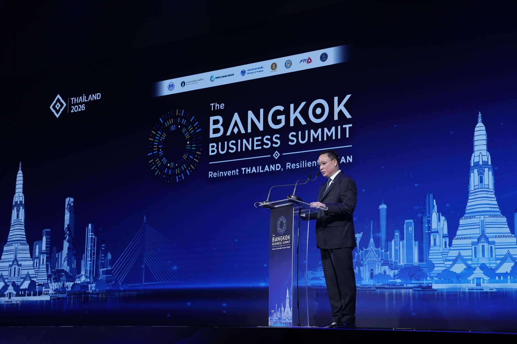 ภาพผู้บริหารระดับสูงกำลังกล่าวสุนทรพจน์บนเวทีงาน The Bangkok Business Summit 2026 โดยมีฉากหลังเป็นภาพกราฟิกสถานที่สำคัญของกรุงเทพมหานคร เช่น วัดอรุณราชวรารามและเสาชิงช้า