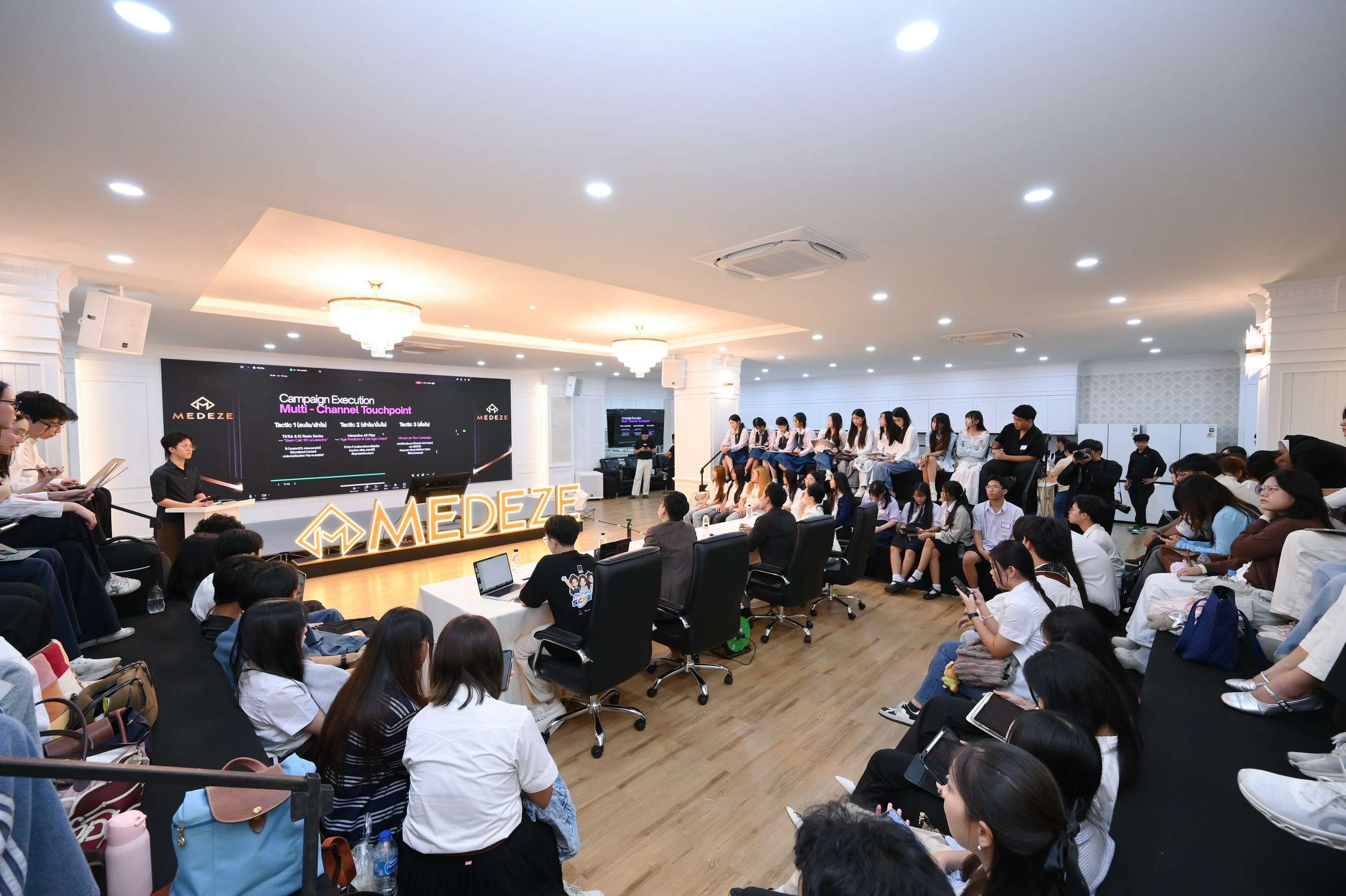 ภาพบรรยากาศกลุ่มนักเรียนและนักศึกษาจำนวนมากกำลังนั่งฟังการนำเสนอแผนธุรกิจอย่างตั้งใจ ภายในห้องจัดกิจกรรมของบริษัท เมดีซ กรุ๊ป จำกัด (มหาชน)