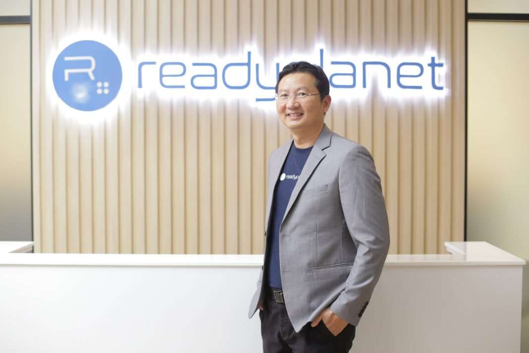 Songyot-Kanthamanon-CEO-Readyplanet-Hotel-Loyalty-Pointspot ภาพนายทรงยศ คันธมานนท์ ประธานเจ้าหน้าที่บริหาร บริษัท เรดดี้แพลนเน็ต จำกัด (มหาชน) หรือ READY สวมชุดสูทสีเทาทับเสื้อยืดสีน้ำเงิน ยืนยิ้มด้วยความมั่นใจบริเวณหน้าเคาน์เตอร์ต้อนรับที่มีป้ายโลโก้ Readyplanet เพื่อประกาศเปิดตัวโซลูชัน Hotel Loyalty Program by Pointspot