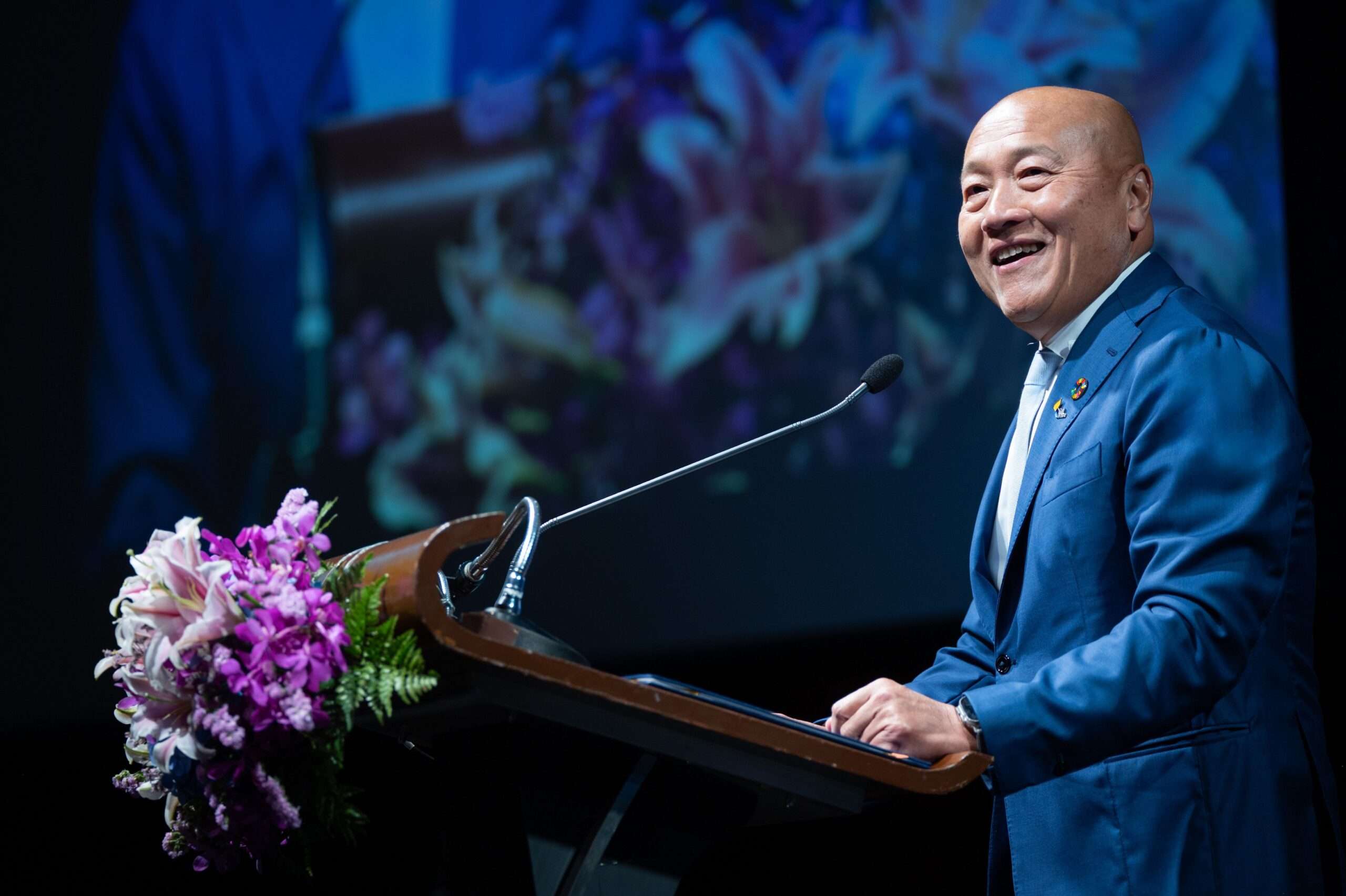ภาพนายธีรพงศ์ จันศิริ ประธานเจ้าหน้าที่บริหาร บริษัท ไทยยูเนี่ยน กรุ๊ป จำกัด (มหาชน) สวมชุดสูทสีน้ำเงิน ยืนยิ้มและกล่าวสุนทรพจน์บนโพเดียมที่มีการตกแต่งด้วยดอกไม้กล้วยไม้ ในงาน Responsible Seafood Summit 2026