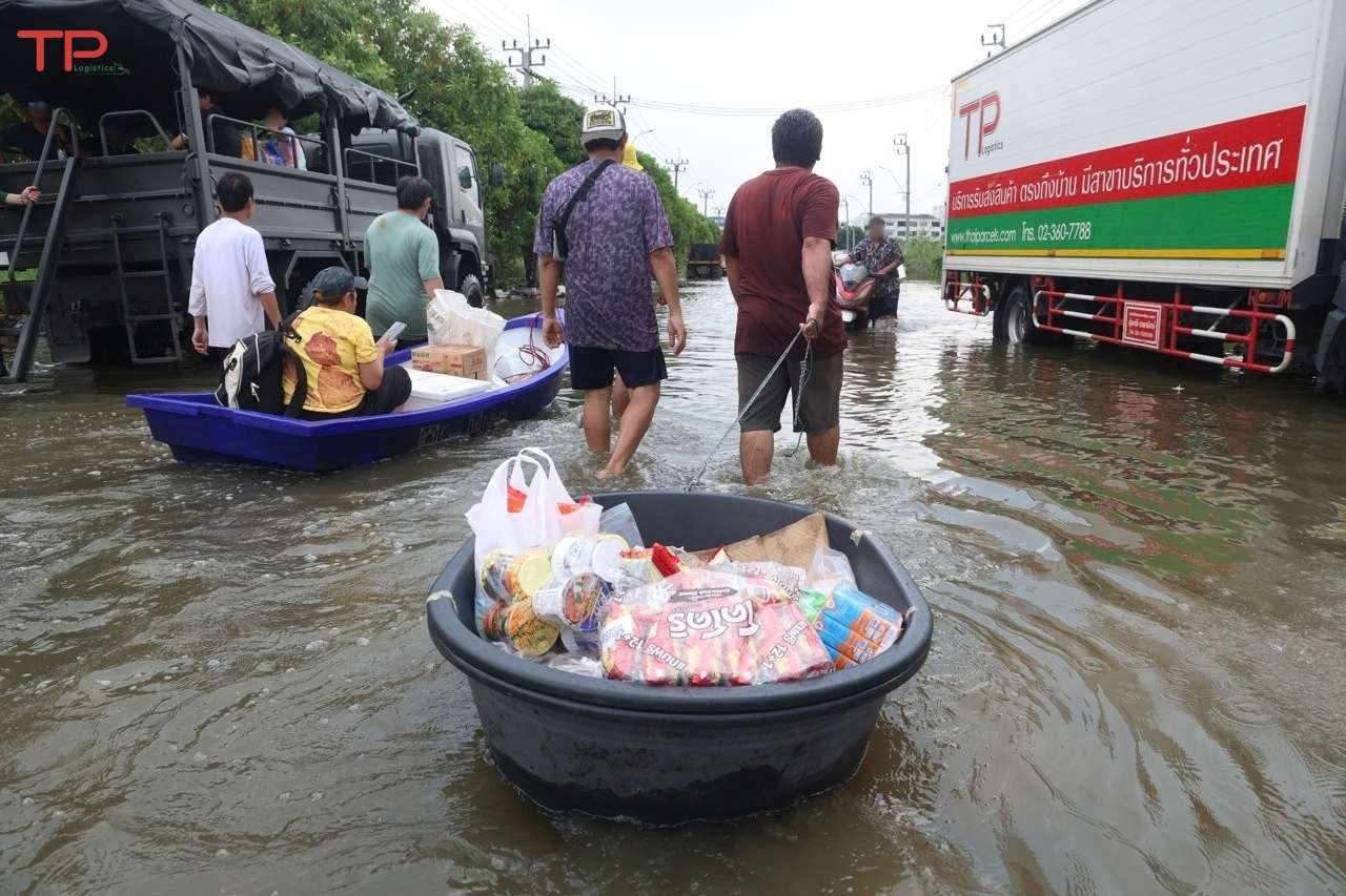 ประชาชนกำลังเดินลุยน้ำท่วมสูงระดับเข่า โดยลากกะละมังสีดำขนาดใหญ่ที่บรรจุบะหมี่กึ่งสำเร็จรูปและอาหารแห้ง เพื่อนำไปแจกจ่ายในพื้นที่ประสบภัย โดยมีรถบรรทุกของ TP Logistics จอดอยู่ด้านข้าง
