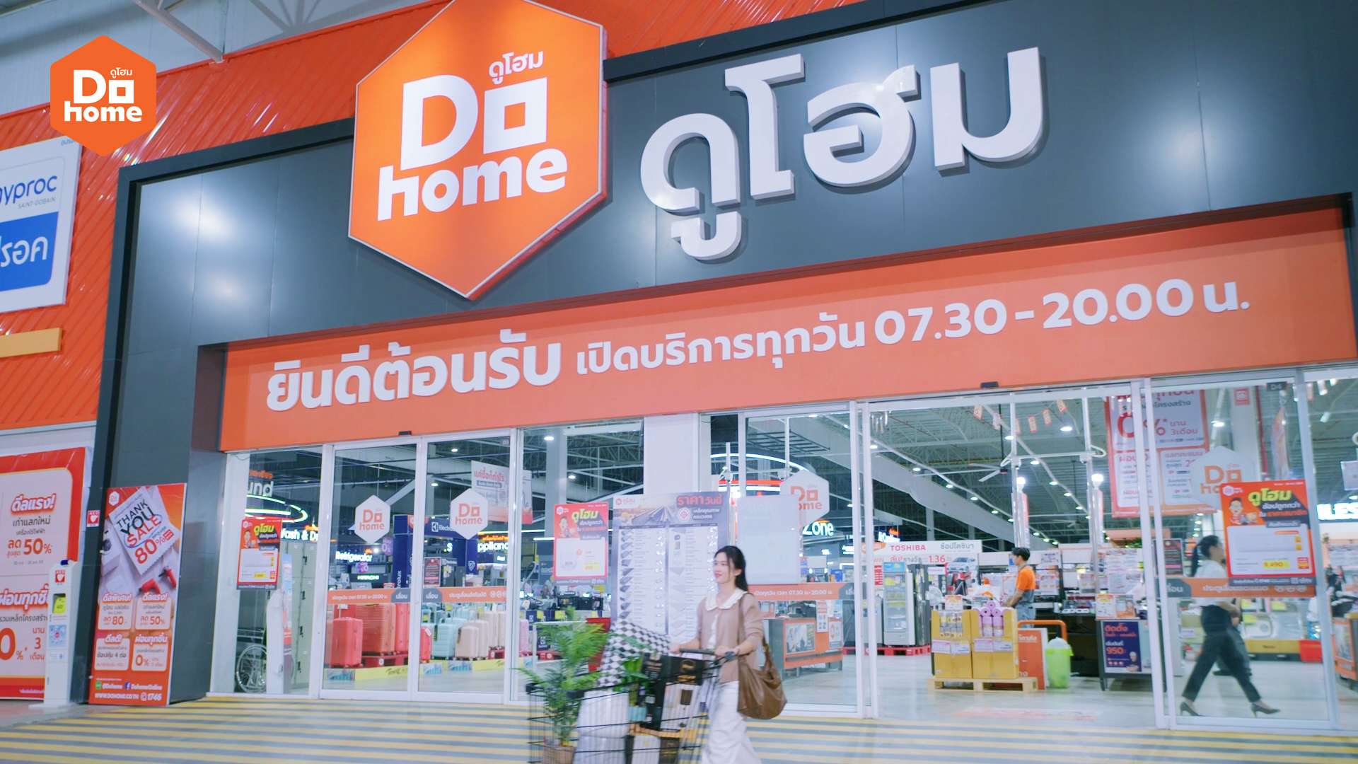 บรรยากาศบริเวณหน้าสาขาของร้านดูโฮม (Dohome) ศูนย์รวมวัสดุก่อสร้างและของตกแต่งบ้าน ป้ายโลโก้สีส้มขนาดใหญ่พร้อมข้อความยินดีต้อนรับเปิดบริการทุกวัน โดยมีลูกค้ายกของใส่รถเข็นเดินออกมา