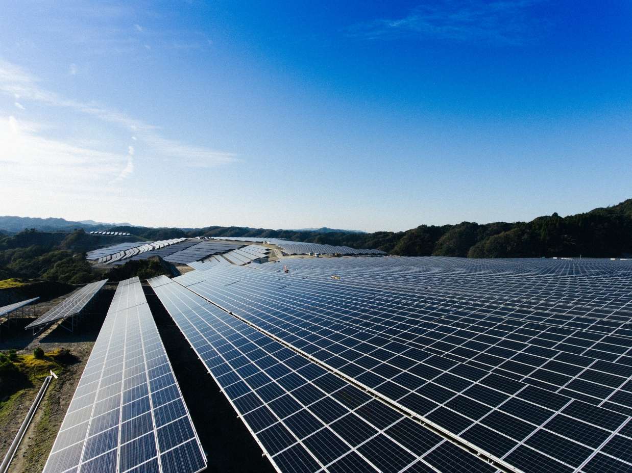 ภาพมุมสูงของโครงการโรงไฟฟ้าพลังงานแสงอาทิตย์ขนาดใหญ่ (Solar Farm) ที่มีแผงโซลาร์เซลล์ติดตั้งเรียงรายอย่างเป็นระเบียบครอบคลุมพื้นที่กว้างขวาง ท่ามกลางธรรมชาติและภูเขา