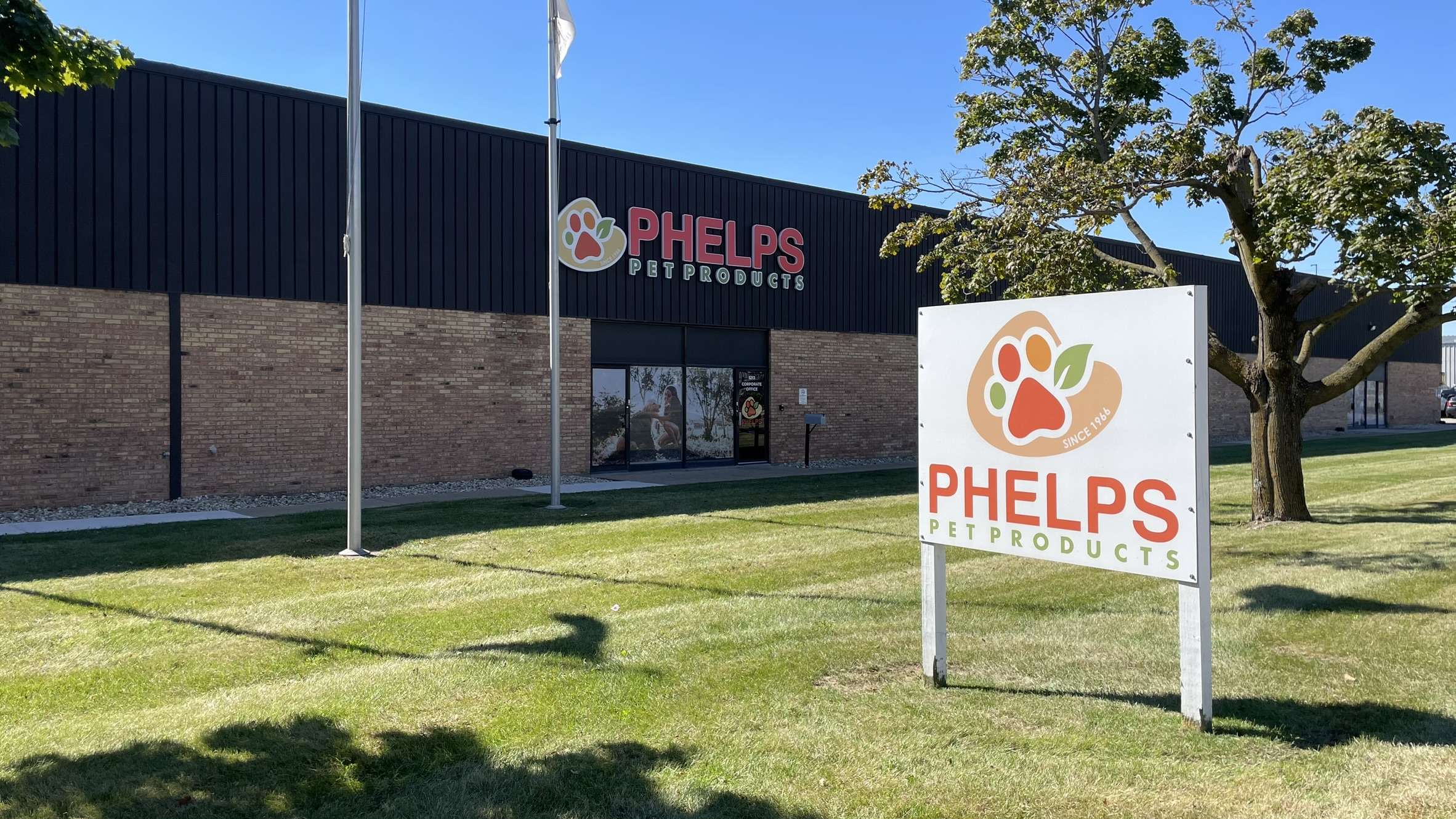 ป้ายชื่อและโลโก้ขนาดใหญ่ของบริษัท Phelps Pet Products ตั้งอยู่บนสนามหญ้าสีเขียว ด้านหน้าอาคารสำนักงานและโรงงานชั้นเดียวที่ก่อด้วยอิฐสีน้ำตาล