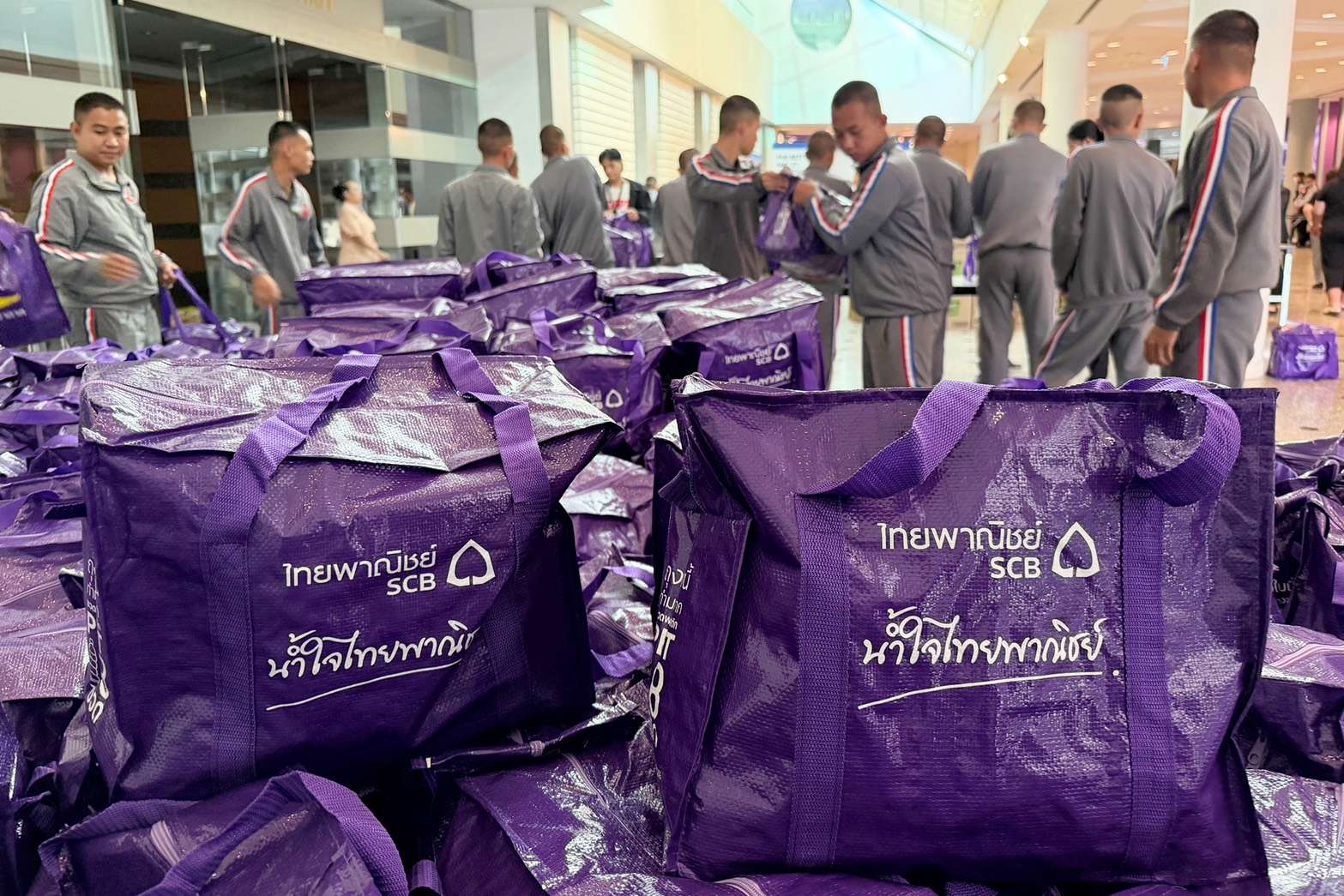 ถุงยังชีพสีม่วงพิมพ์ข้อความ น้ำใจไทยพาณิชย์ จำนวนมากวางเรียงราย โดยมีเจ้าหน้าที่ทหารกำลังช่วยกันจัดเตรียมและลำเลียงอยู่บริเวณด้านหลัง