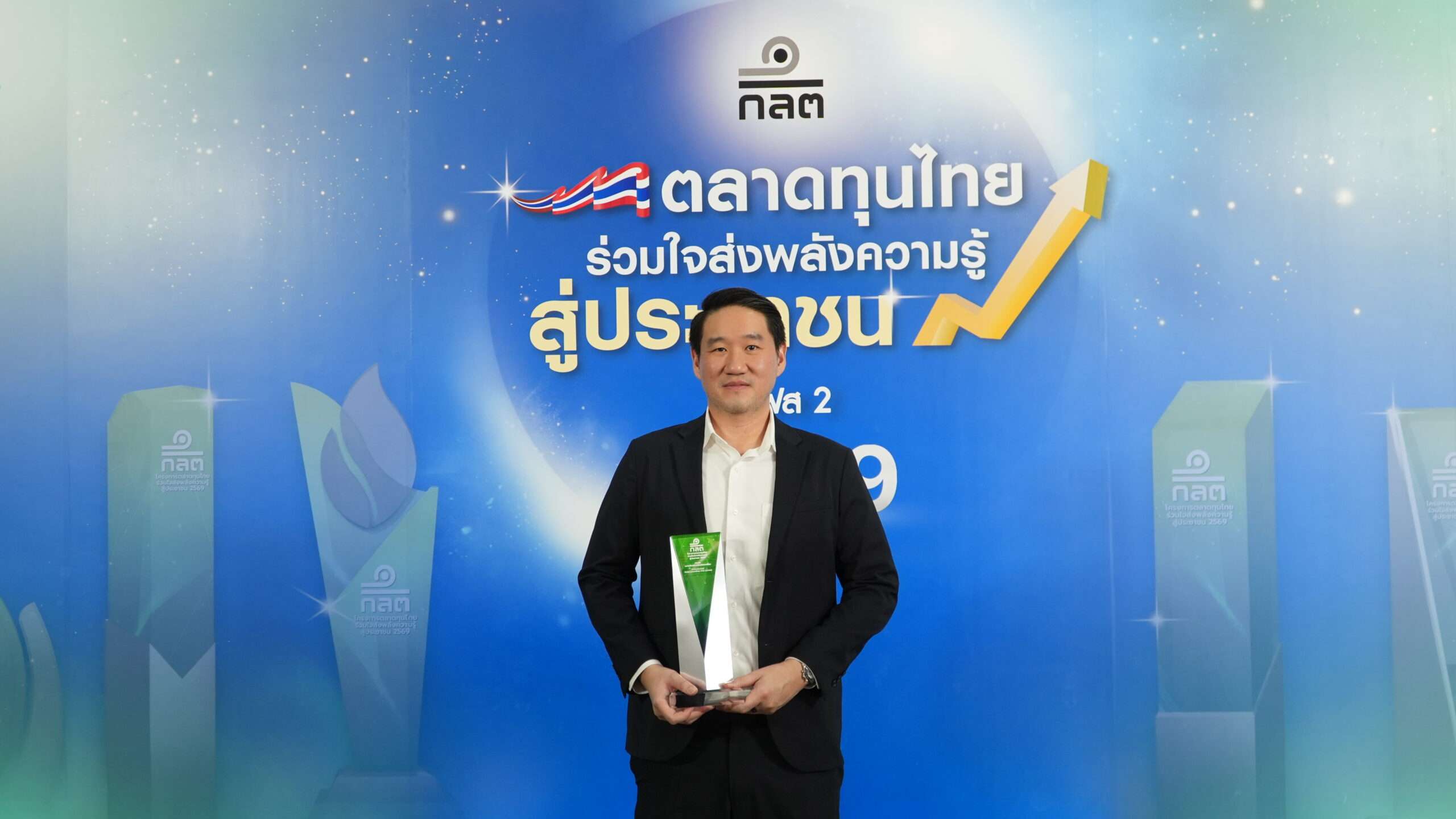 นายสานุพงศ์ สุทัศน์ธรรมกุล กรรมการผู้จัดการ บล.ฟิลลิป ถือถ้วยรางวัลความคิดสร้างสรรค์ยอดเยี่ยม (Creativity Award) ยืนยิ้มหน้าป้ายงาน ตลาดทุนไทย ร่วมใจส่งพลังความรู้ สู่ประชาชน เฟส 2