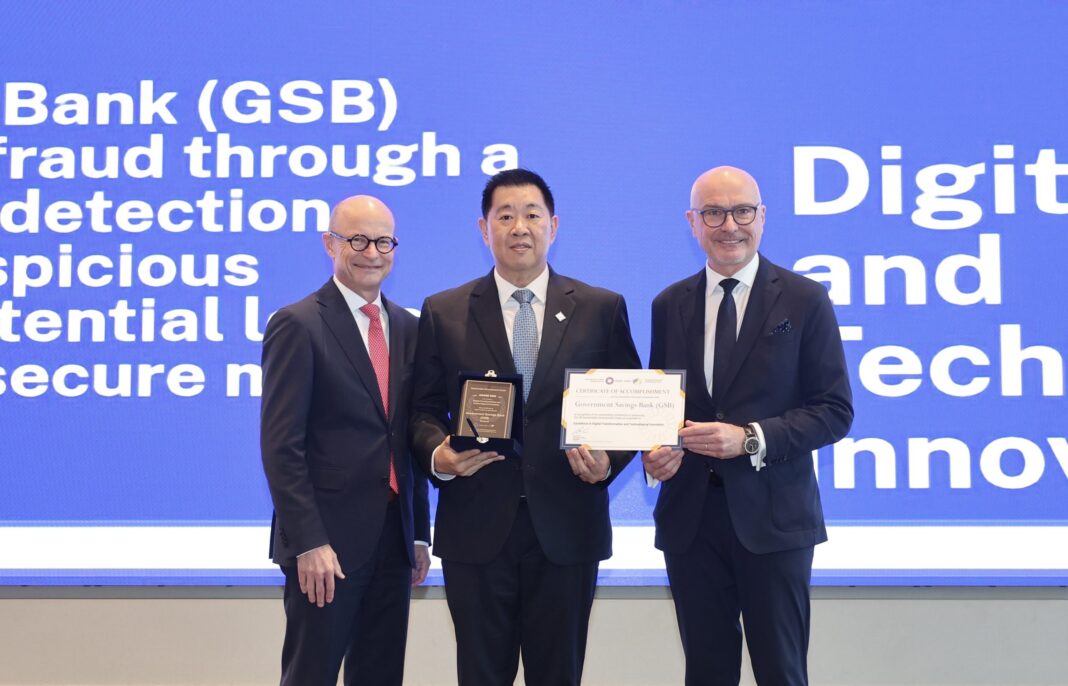 Songpol-Chewapunyaroj-GSB-WSBI-ESBG-SDG-Awards-2026 นายทรงพล ชีวะปัญญาโรจน์ ผู้อำนวยการธนาคารออมสิน ยืนตรงกลางรับมอบโล่รางวัลและประกาศนียบัตร WSBI-ESBG SDG Awards 2026 สาขา Digital Transformation and Technological Innovation ร่วมกับผู้บริหารระดับสูงชาวต่างชาติ บนเวทีที่มีฉากหลังหน้าจอสีน้ำเงินระบุชื่อรางวัล
