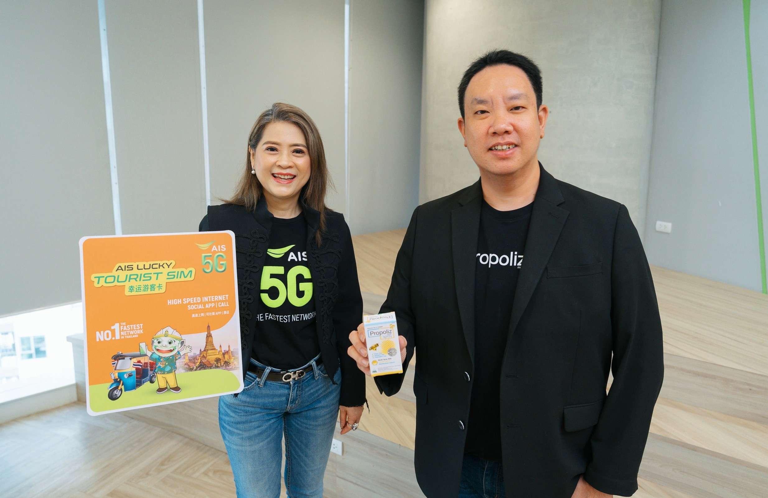 ผู้บริหารหญิงจาก AIS ถือป้ายโฆษณาจำลอง AIS LUCKY TOURIST SIM ยืนคู่กับนายธนัท พลอยดนัย ผู้อำนวยการฝ่ายการตลาด TMAN ที่กำลังถือผลิตภัณฑ์ Propoliz Mouth Spray เพื่อโปรโมตความร่วมมือในแคมเปญใหม่