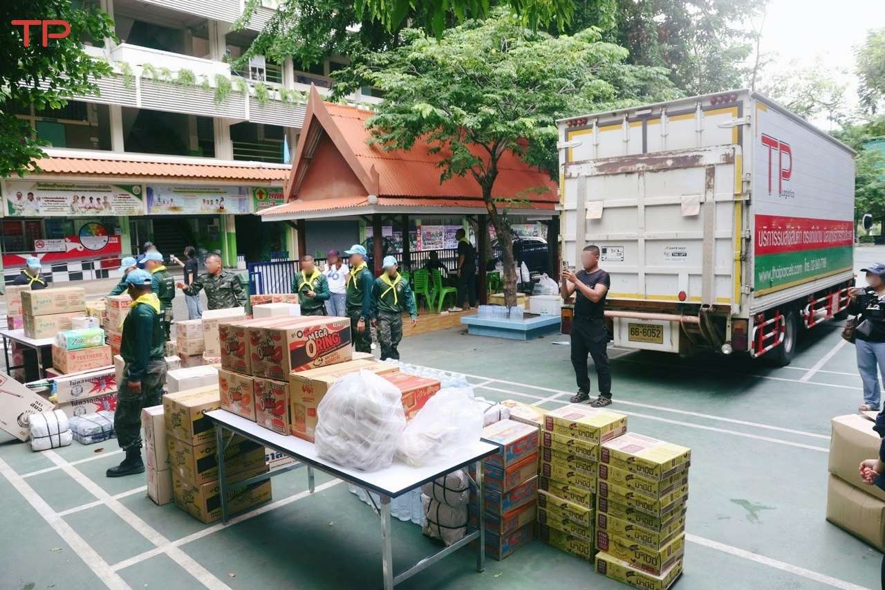 รถบรรทุกของ TP Logistics จอดเทียบจุดรับบริจาค โดยมีเจ้าหน้าที่ทหารและจิตอาสาช่วยกันจัดเตรียมและลำเลียงกล่องบรรจุสิ่งของอุปโภคบริโภค น้ำดื่ม และอาหารแห้งจำนวนมากที่ลานอเนกประสงค์