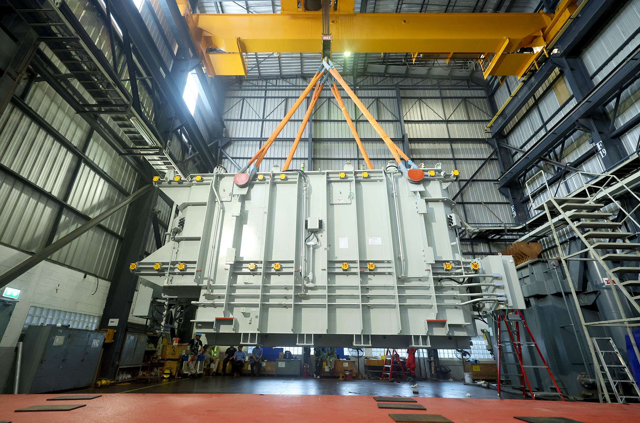หม้อแปลงไฟฟ้ากำลัง (Power Transformer) ขนาดใหญ่พิเศษถูกเครนยกเคลื่อนย้ายภายในอาคารโรงงานผลิตของ TRT โดยมีเจ้าหน้าที่วิศวกรสวมหมวกนิรภัยคอยดูแลความปลอดภัยอยู่ด้านล่าง