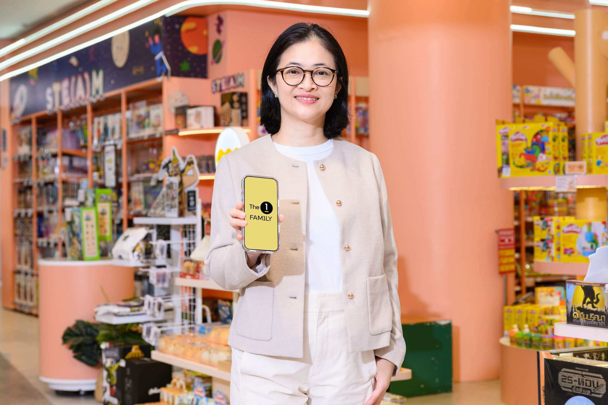 คุณทิพาภรณ์ ประสมทรัพย์ Head of Loyalty Program and CRM จาก The 1 Central Group ถือสมาร์ทโฟนที่แสดงหน้าจอแอปพลิเคชัน The 1 ยืนยิ้มอยู่บริเวณโซนของเล่นเด็ก