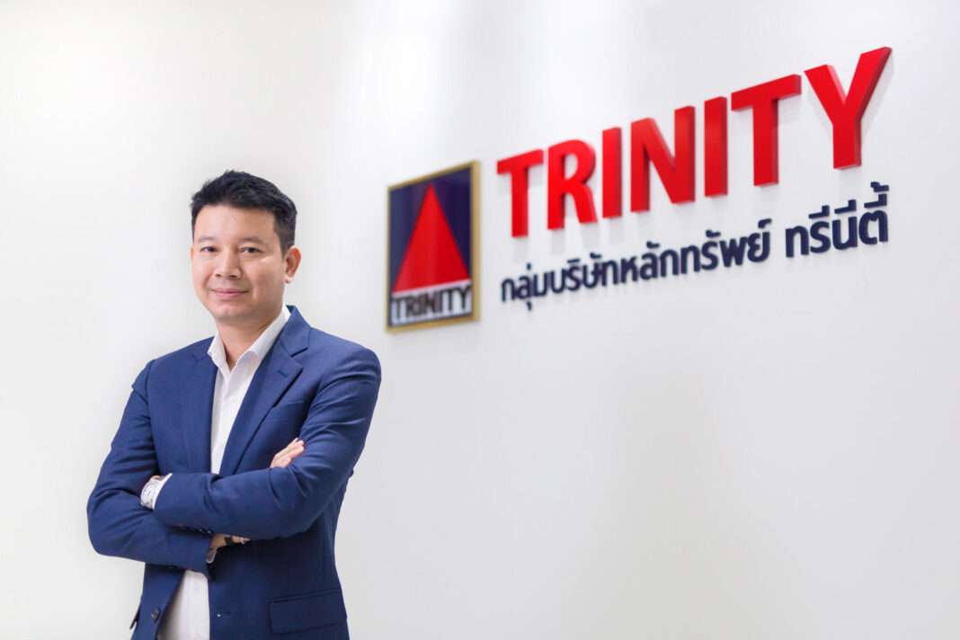 Weerapat-Petchkupt-CEO-Trinity-Securities-Hackathon ดร.วีรพัฒน์ เพชรคุปต์ ประธานเจ้าหน้าที่บริหาร บริษัทหลักทรัพย์ ทรีนีตี้ จำกัด สวมเสื้อสูทสีน้ำเงินทับเสื้อเชิ้ตสีขาว ยืนกอดอกด้วยความมั่นใจบริเวณหน้าโลโก้กลุ่มบริษัทหลักทรัพย์ ทรีนีตี้