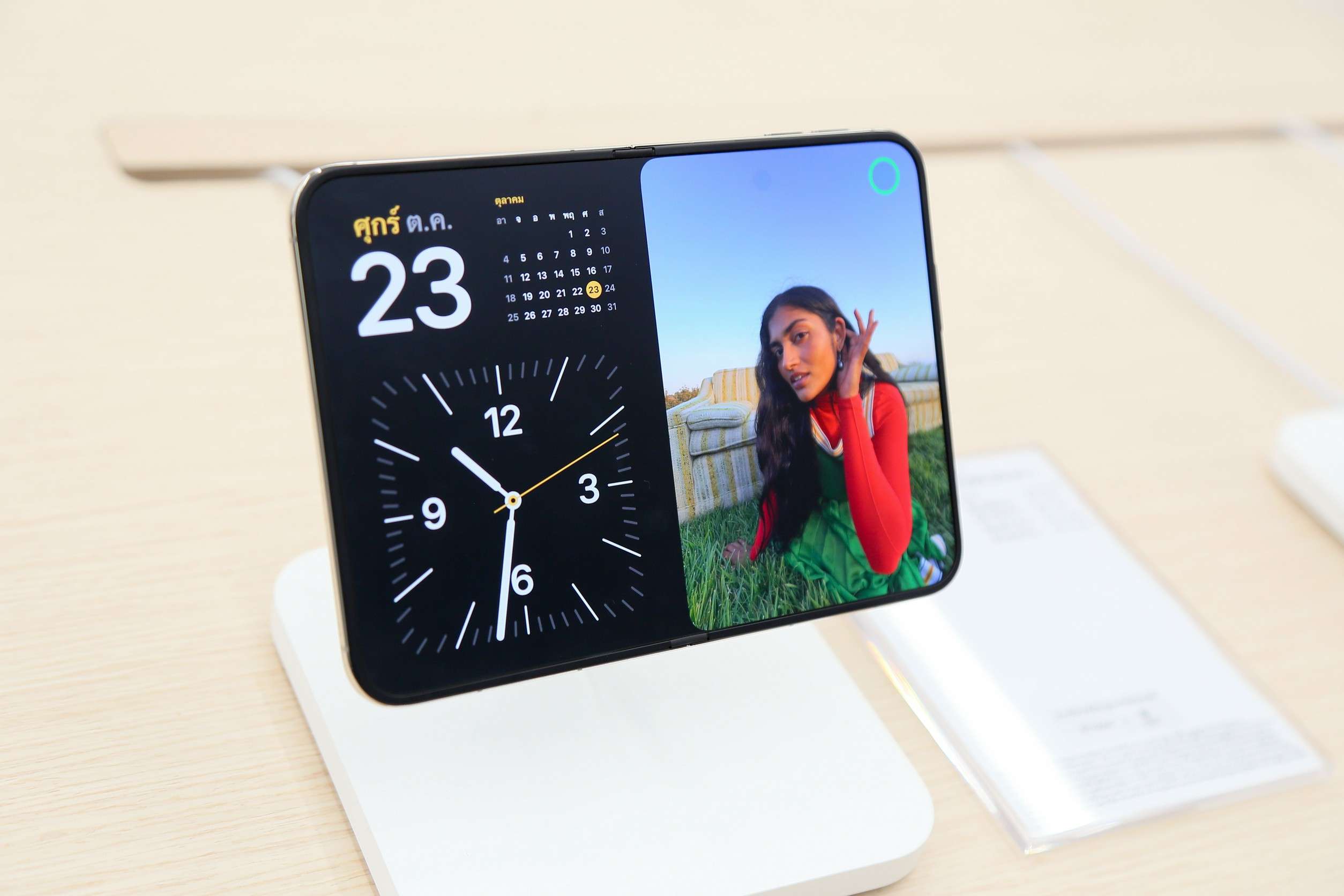 ภาพตัวเครื่อง iPhone Duo สมาร์ตโฟนจอพับรุ่นแรกของ Apple กางออกตั้งโชว์บนแท่นชาร์จไร้สาย หน้าจอแสดงผลแบบแบ่งครึ่ง ด้านซ้ายเป็นนาฬิกาและปฏิทิน ด้านขวาเป็นรูปภาพนางแบบ ภายในร้าน iStudio